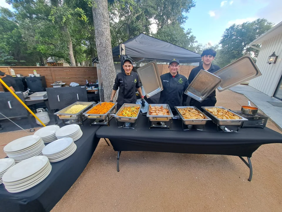 Three Mana Sabroso team members behind an outdoor buffet with serving trays ready