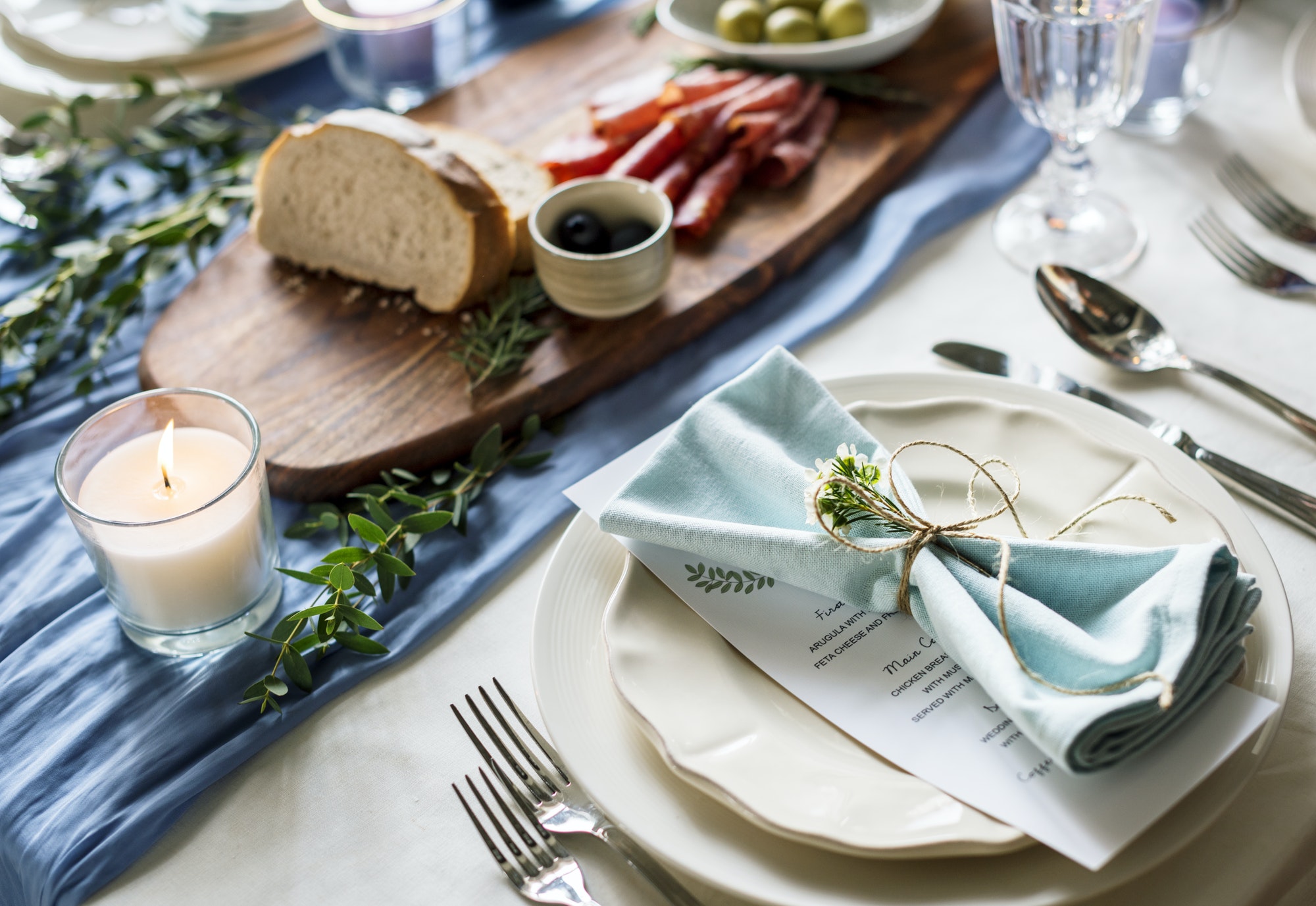 A charcuterie board with rolls of meat, olives, and bread next to silverware and a plat with an elegantly folded napkin