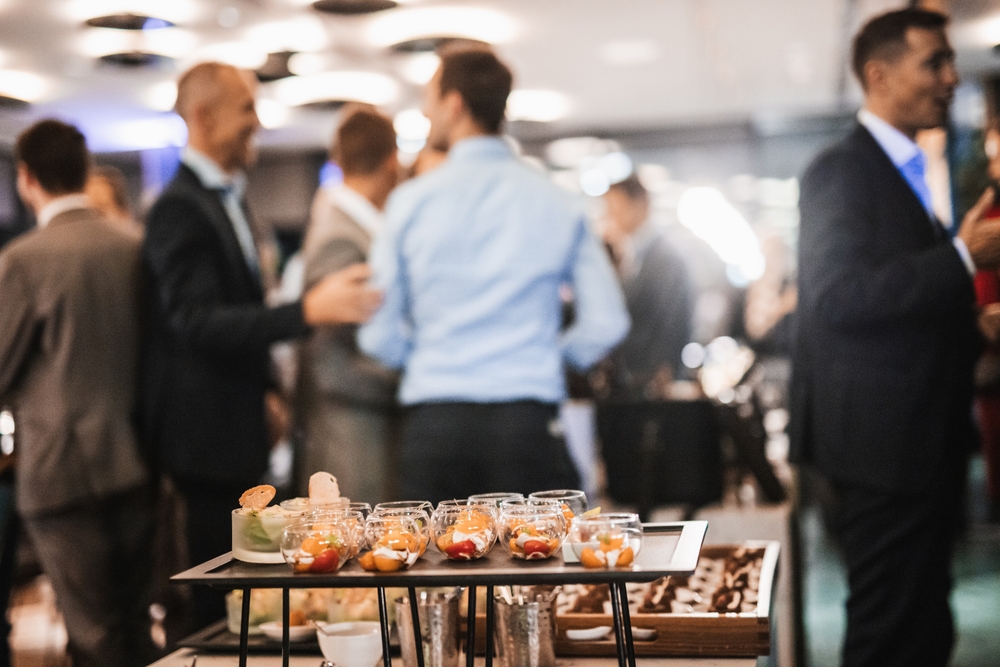 Close-up of a catering table covered with small, carefully prepared portions in the foreground while people talk in the background