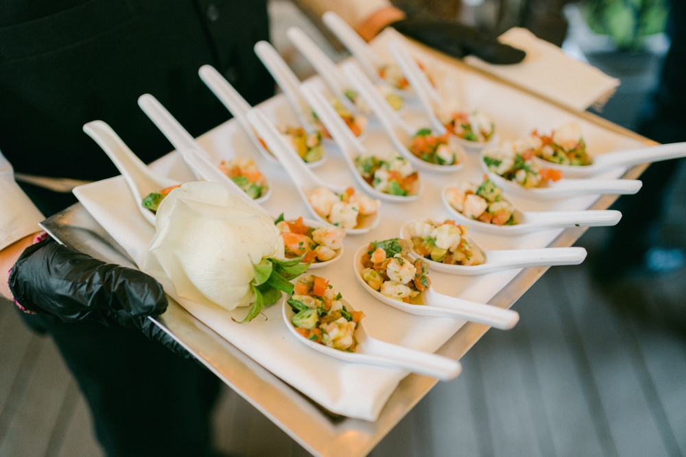 Gloved hands holding a tray of white spoons, each filled with a collection of seafood and vegetables