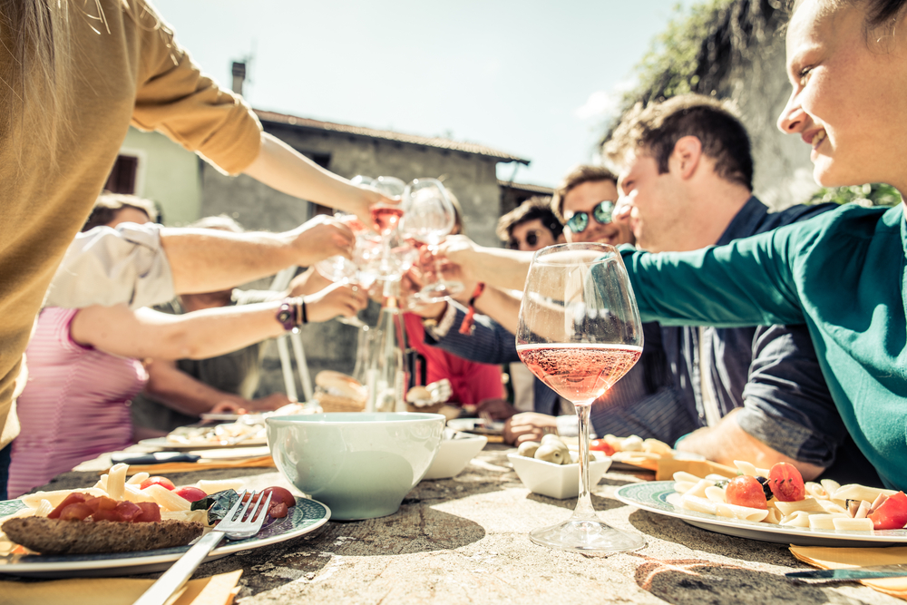 A group of people sitting at a table outside of a home eating, clinking their glasses together